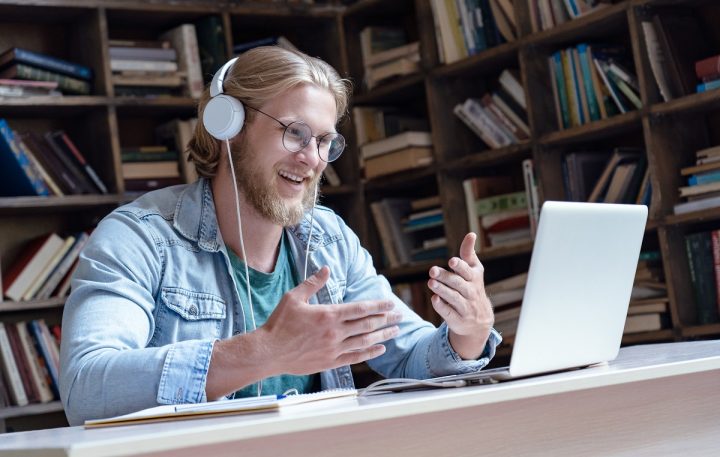 Young male teacher with headphones on at desk with computer. Bookshelf in the background