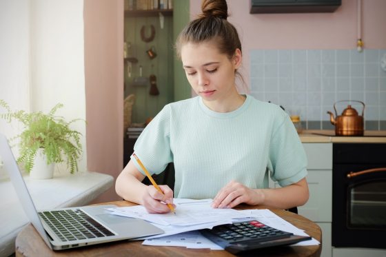 Teenage girl at kitchen table doing homework with computer, calculator and papers