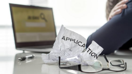 Person with head in hands in front of computer searching for jobs. Crumpled piece of paper with the word "application" next to the person.