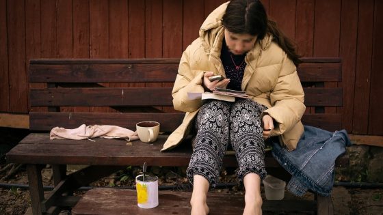 Young girl with school book in lap sitting outside on wooden bench barefoot