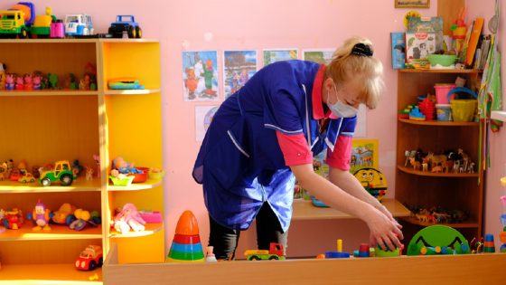 Child care worker wearing face mask tidying up toys in play room