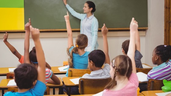 Diverse students in classroom raising their hands. Teacher standing in front of a blackboard
