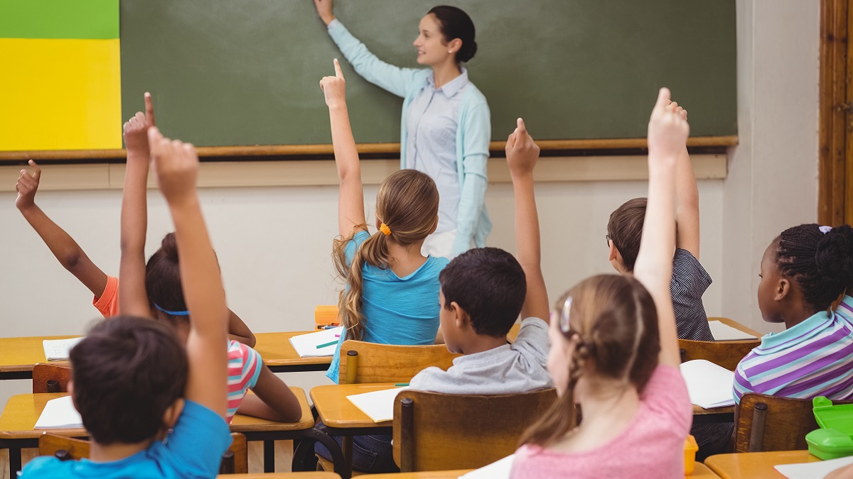 Diverse students in classroom raising their hands. Teacher standing in front of a blackboard