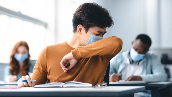 High school student sitting at desk in school, wearing a face mask and coughing into his elbow
