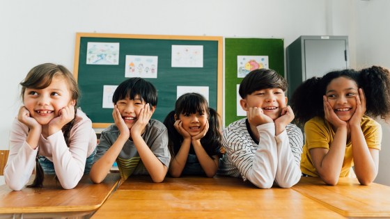 Diverse school children smiling as they lean over their desks in the classroom