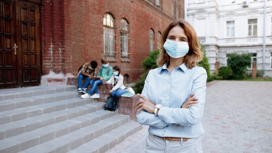 Female school principal wearing face mask with arms crossed standing outside school building with children sitting behind