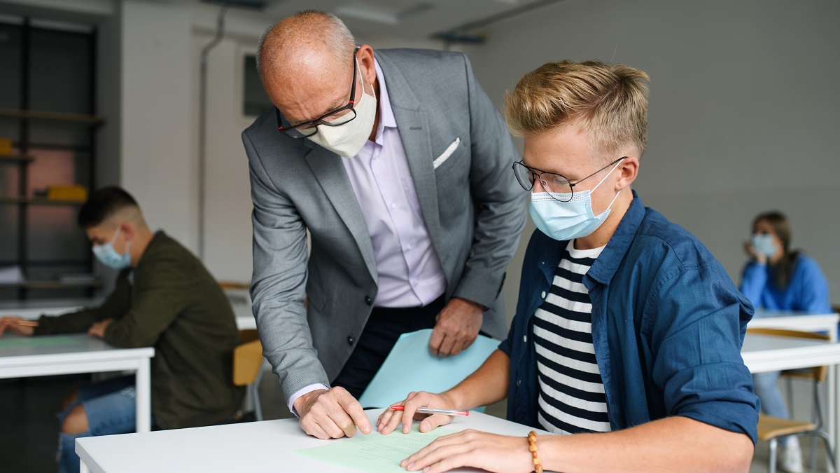 Older male teacher wearing face mask helping high school student with his work at the student's desk