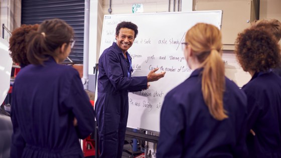 Mechanic vocational teacher in front of whiteboard in mechanic overalls surrounded by students listening