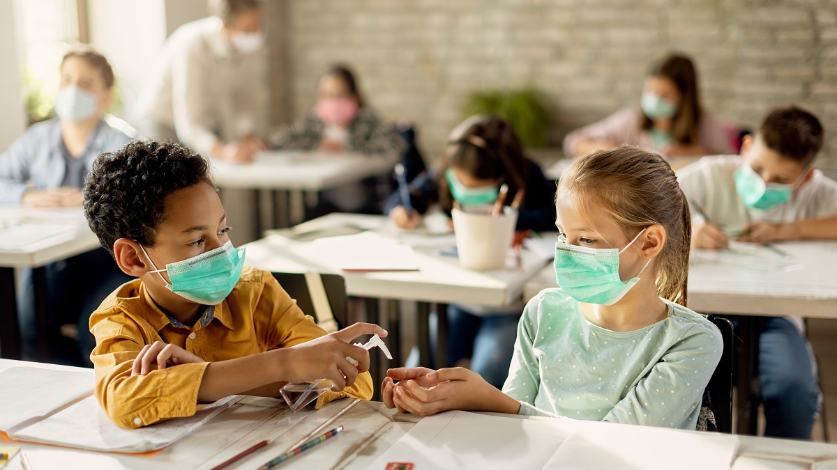 Male school student sprays female student's hands with sanitiser. All students in the classroom are wearing face masks.
