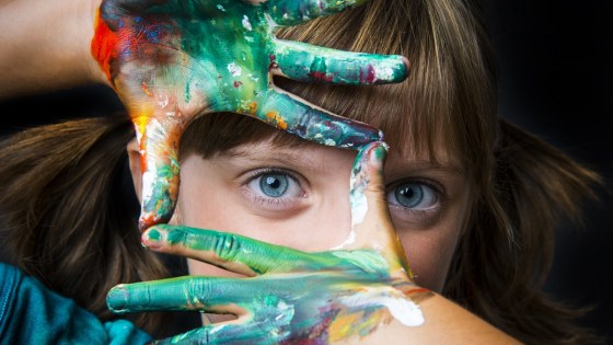 Young girl holding her fingers in the shape of a square in front of her face and looking through them with one eye. Her hands are covered in paint.