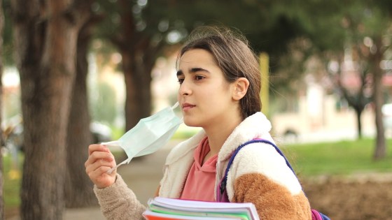 Young female school student walking outside with school books in her arms, taking off covid face mask