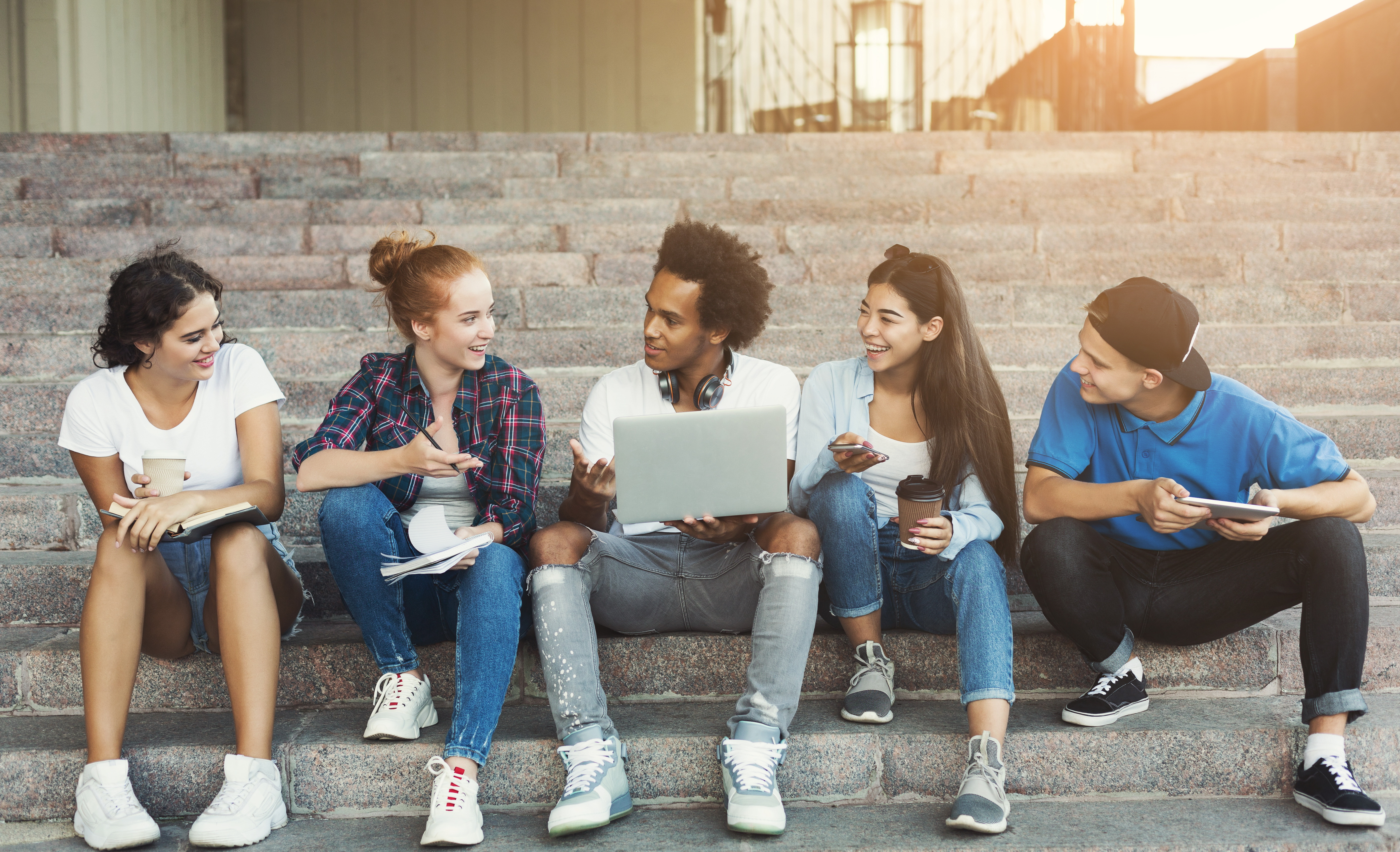 5 young people sit on some concrete steps discussion something with each other. The boy in the middle holds a laptop. The sun is setting