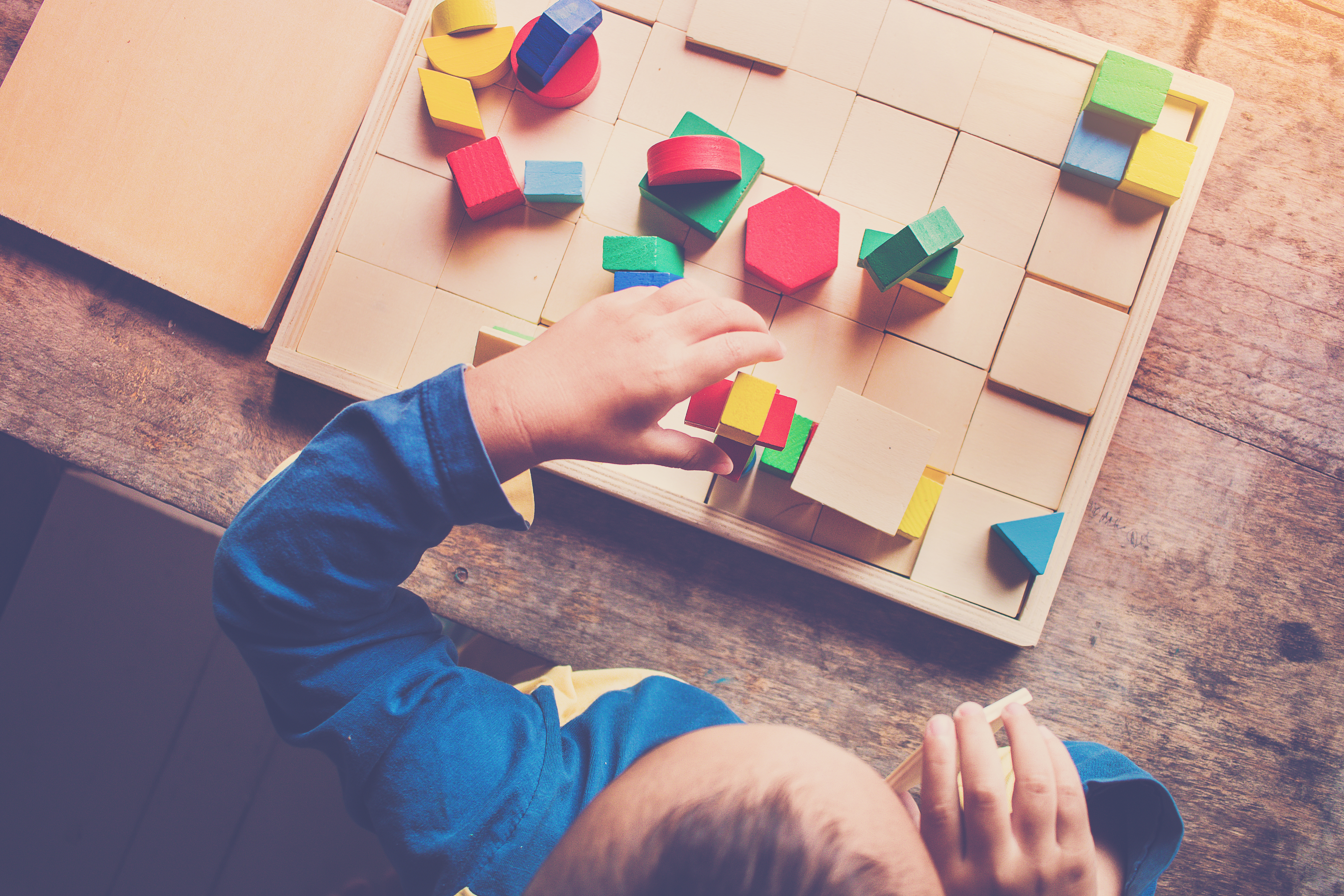 Young boy playing with multiple coloured blocks on a wooden table. Pictured from above. Boy is grabbing the block piled up in front of him