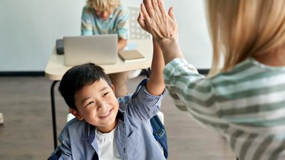 a young asian boy is seated and high-fiving his teacher from his desk in a classroom